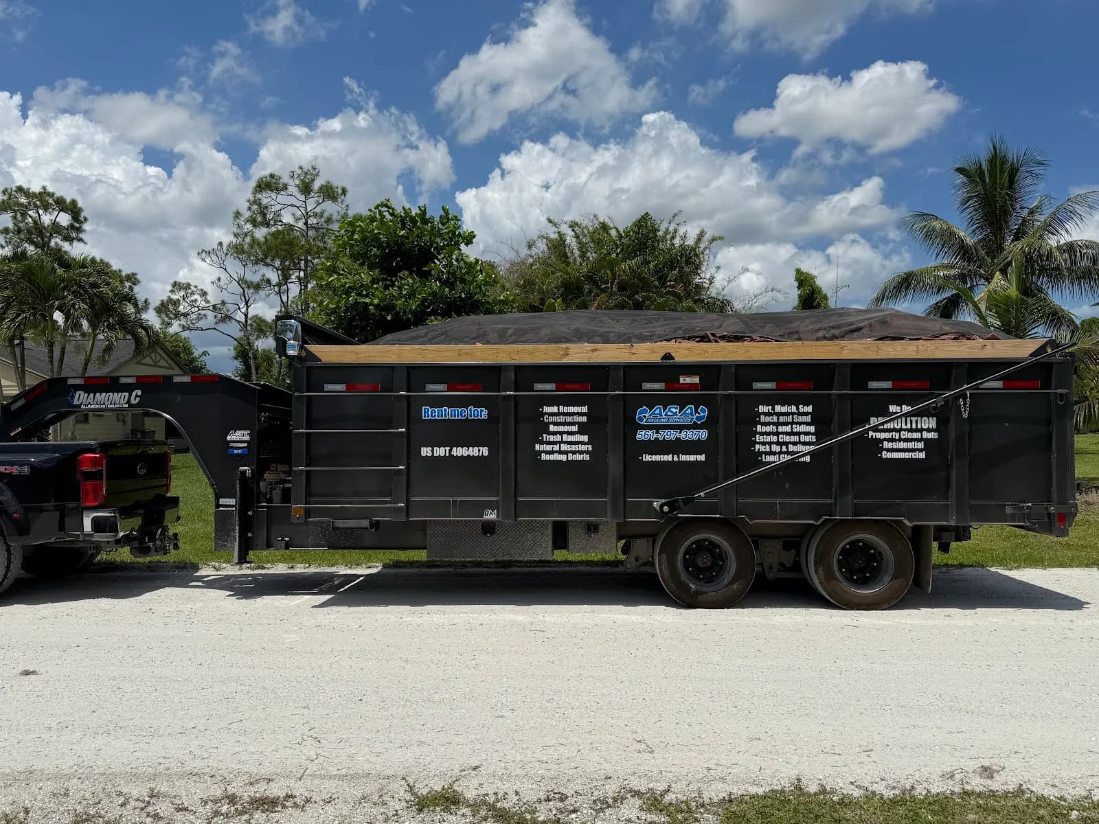 Black gooseneck dump trailer full of roofing debris under palm trees and a blue sky