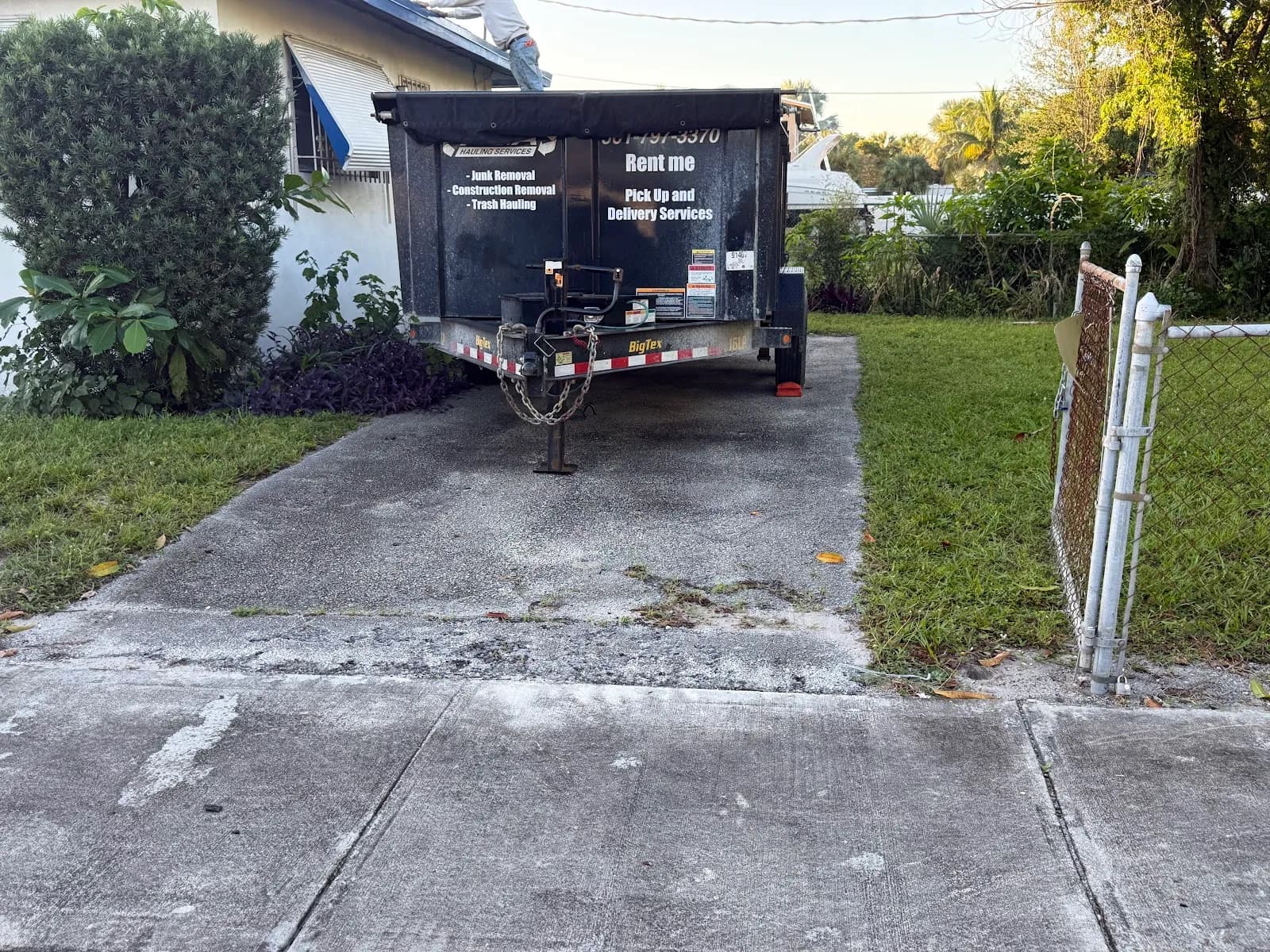 Black dump trailer with signage listing junk removal, construction removal, trash hauling, dump trailer rental, pick up and delivery services, and the phone number